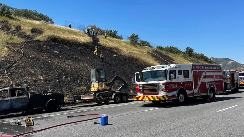 A burned pickup truck sits on the side of the road next to a Park City Fire District truck as firefighters work on the burned hillside.