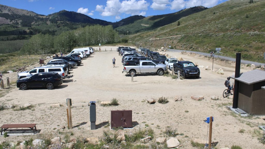Cars fill a dirt parking lot with lush green mountains in the background.