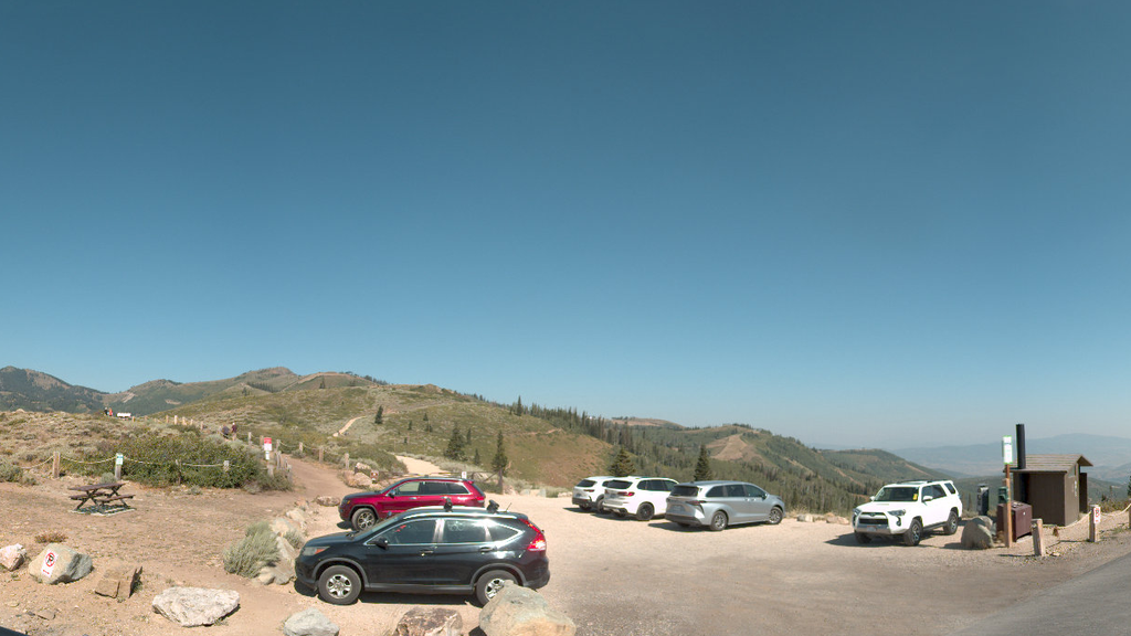 A panoramic view of a dirt parking area with six cars surrounded by a picnic table, a short trail leading to an overlook, a trail leading into the mountains, and a pit toilet.
